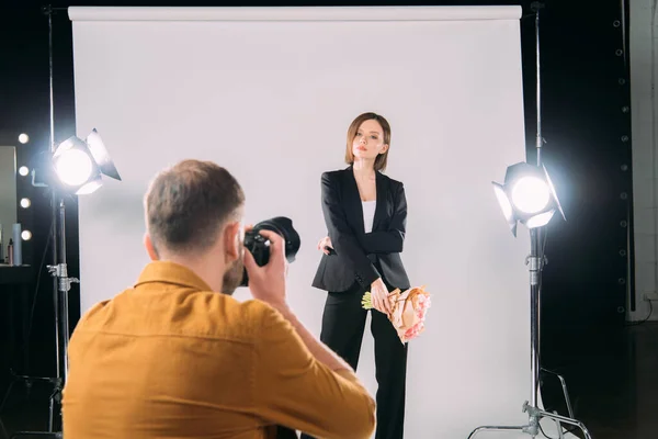 Selective focus of stylish model with bouquet posing at photographer with digital camera in photo studio — Stock Photo