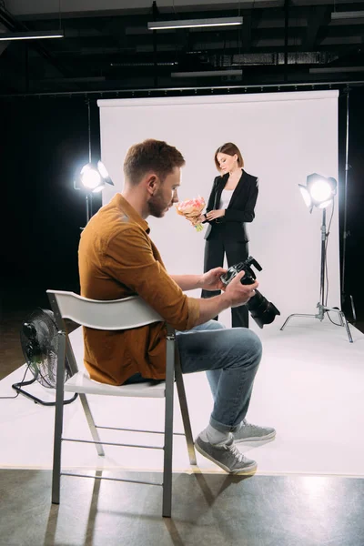 Handsome photographer holding digital camera near elegant model holding bouquet of flowers in photo studio — Stock Photo