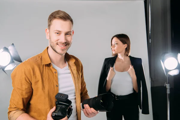 Selective focus of handsome photographer smiling while holding lens and digital camera near beautiful model in photo studio — Stock Photo