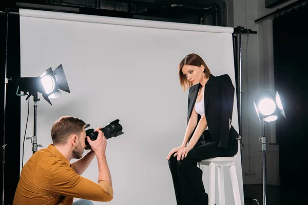 Vista laterale del fotografo che lavora con elegante modello seduto sulla sedia in studio fotografico — Foto stock