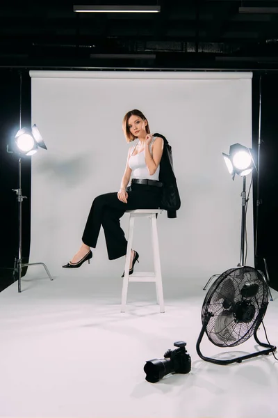 Side view of beautiful model holding jacket while sitting on chair near electric fan and digital camera in photo studio — Stock Photo