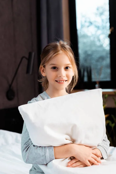 Cheerful kid smiling while holding pillow in bedroom — Stock Photo