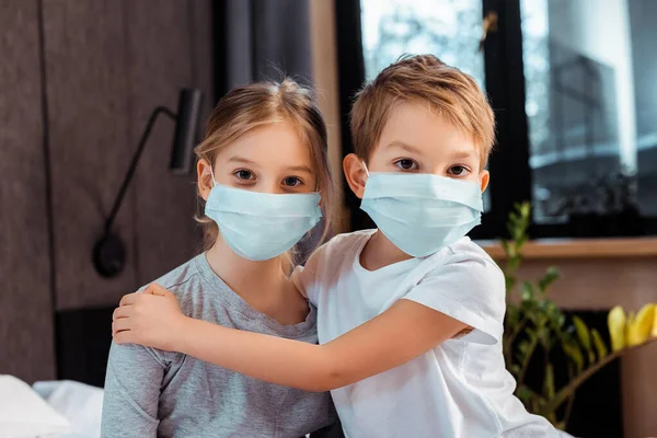 Cute brother hugging sister in medical mask at home — Stock Photo