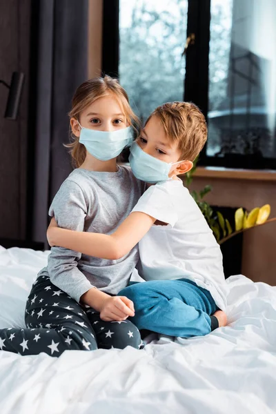 Cute brother hugging sister in medical mask while sitting on bed — Stock Photo