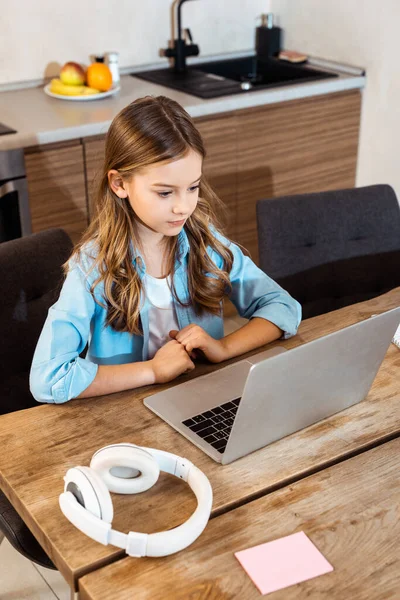 Lindo niño en línea estudiando mientras mira el portátil cerca de los auriculares en casa - foto de stock