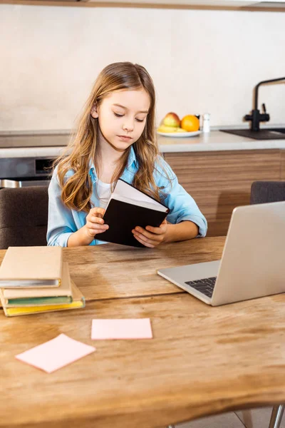 Foyer sélectif d'enfant tenant le livre près de l'ordinateur portable tout en étudiant en ligne à la maison — Photo de stock