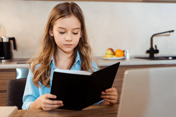 Foyer sélectif de livre de lecture mignon enfant près d'un ordinateur portable tout en e-learning à la maison — Photo de stock