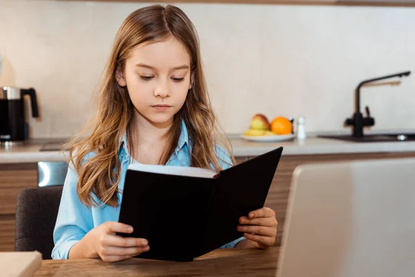 Enfoque selectivo del libro lindo de la lectura del cabrito cerca del ordenador portátil mientras que e-learning en el país - foto de stock