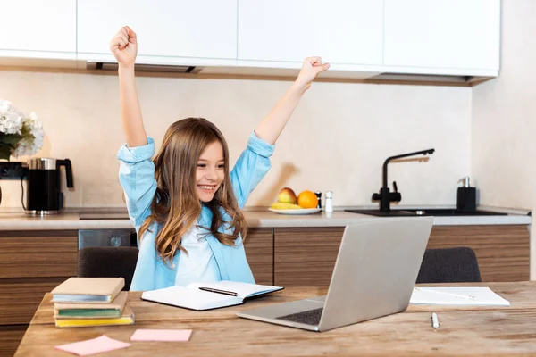 Selective focus of happy kid with hands above head looking at laptop while e-learning at home — Stock Photo