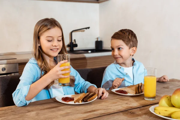 Foco seletivo de criança feliz segurando vidro de suco de laranja perto do irmão e café da manhã saboroso — Fotografia de Stock