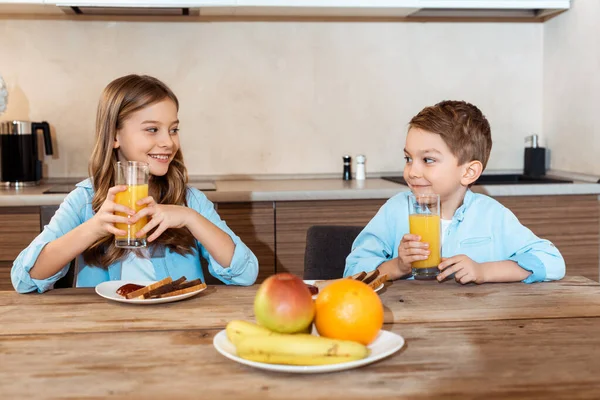 Selektiver Fokus glücklicher Geschwister, die einander anschauen und in der Nähe von leckerem Frühstück Gläser mit Orangensaft halten — Stockfoto