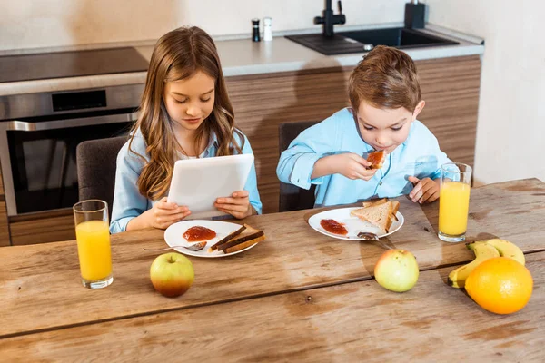 Garçon manger pain grillé avec confiture près de soeur en utilisant une tablette numérique — Photo de stock