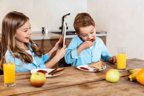 Happy kid using digital tablet near brother eating toast bread with jam — Stock Photo