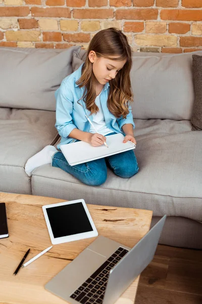 Glückliches Kind sitzt auf Sofa und schreibt in Notizbuch in der Nähe von Gadgets — Stockfoto