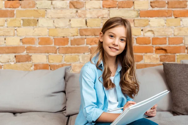 Happy kid sitting on sofa and writing in notebook — Stock Photo