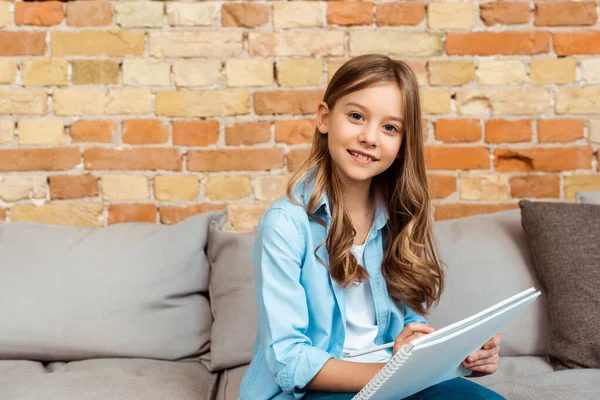 Niño feliz sentado en el sofá y la escritura en el cuaderno - foto de stock