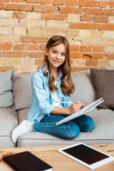 Счастливый ребенок сидит на диване и писать в ноутбуке рядом с цифровой планшет с пустым экраном — стоковое фото