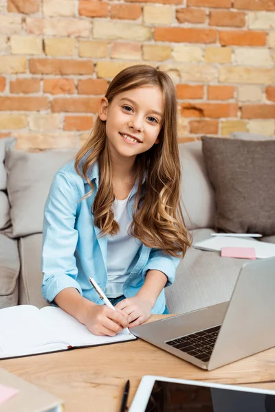 Glückliches Kind schreibt im Notizbuch, während es zu Hause E-Learning macht — Stock Photo