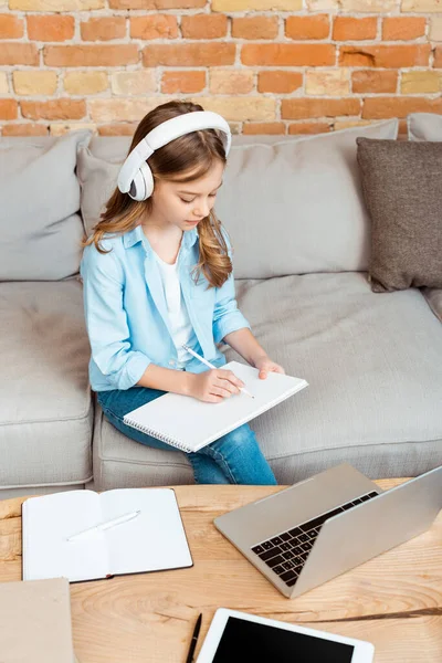 Cute kid in headphones writing in notebook near gadgets — Stock Photo