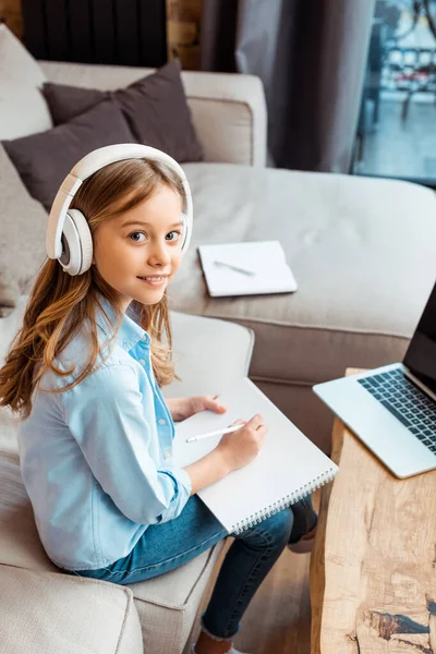 Happy kid in wireless headphones writing in notebook near laptop with blank screen while e-learning at home — Stock Photo