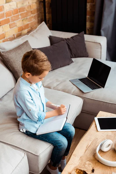 Cute boy writing in notebook near gadgets with blank screen while e-learning at home — Stock Photo