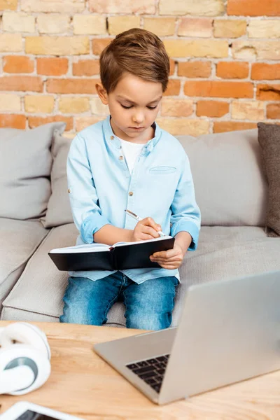 Niño escribiendo en un cuaderno cerca de la computadora portátil mientras e-learning en casa - foto de stock