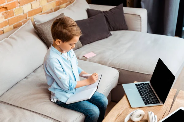 Adorable boy writing in notebook near gadgets with blank screen while e-learning at home — Stock Photo