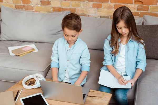 Frères et sœurs mignons en ligne étudier près de gadgets et écouteurs à la maison — Photo de stock