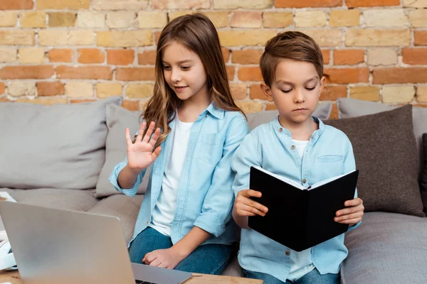 Lindo niño saludando mano mientras tiene videollamada cerca de hermano en sala de estar - foto de stock