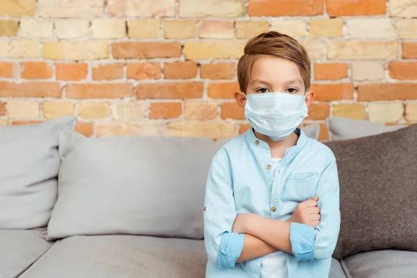 Menino em máscara médica com braços cruzados perto da parede de tijolo na sala de estar — Fotografia de Stock