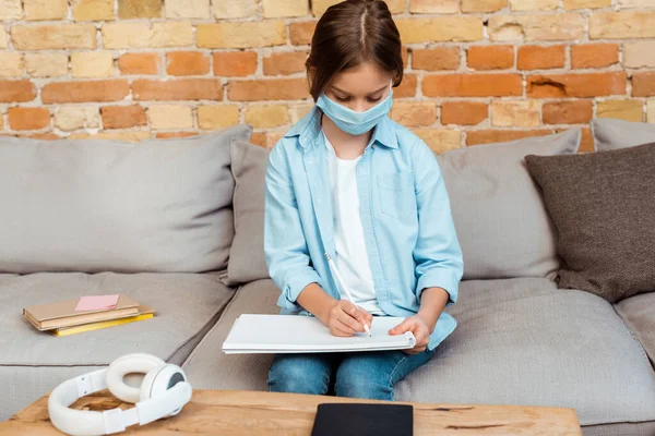 Niño en la escritura de máscara médica en el cuaderno en casa - foto de stock