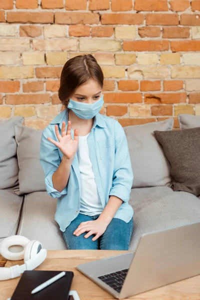 Niño en máscara médica agitando la mano cerca de la computadora portátil - foto de stock