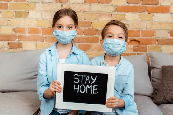 Frères et sœurs dans des masques médicaux tenant un tableau noir avec lettrage à la maison — Photo de stock