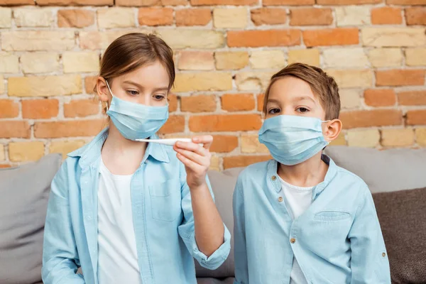 Niño enfermo en máscara médica sosteniendo termómetro digital cerca de hermano - foto de stock