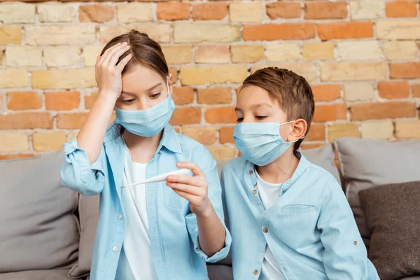 Diseased kid in medical mask holding digital thermometer near brother — Stock Photo