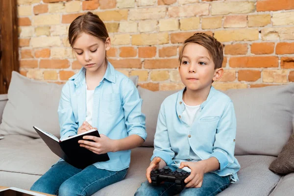 KYIV, UKRAINE - APRIL 27, 2020: sister writing in notebook near brother playing video game — Stock Photo