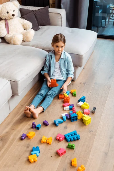 Lindo niño sentado en el piso cerca de bloques de construcción y sofá - foto de stock