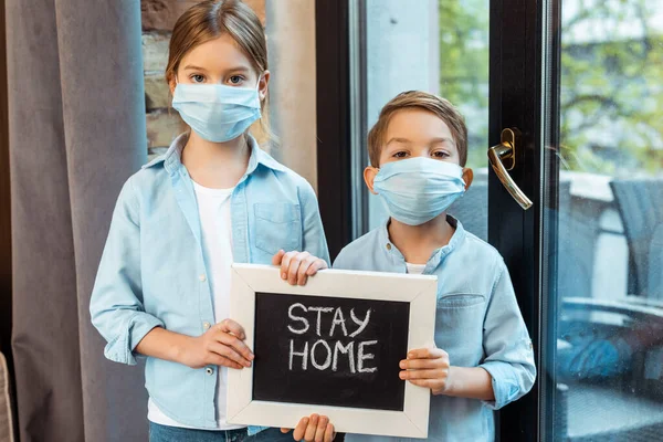 Hermana y hermano en máscaras médicas sosteniendo pizarra con letras de la estancia en casa - foto de stock