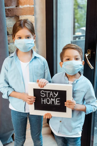 Irmãos em máscaras médicas segurando quadro com ficar em casa lettering — Fotografia de Stock