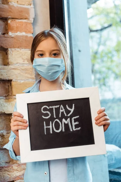 Bambino in maschera medica che guarda la macchina fotografica e tiene la lavagna con scritte a casa — Foto stock