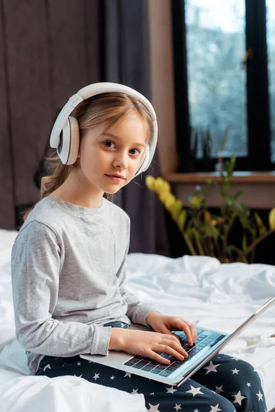 Niño en auriculares inalámbricos usando portátil con e-learning en el dormitorio - foto de stock