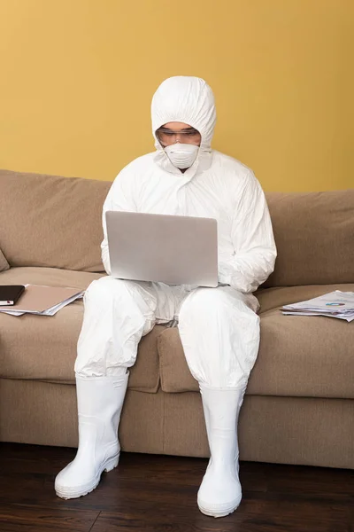 Homem de terno hazmat, máscara de segurança e óculos usando laptop perto de documentos no sofá — Fotografia de Stock