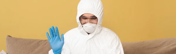 Orientamento panoramico dell'uomo in tuta hazmat, maschera medica e guanto di lattice agitando mano a casa — Foto stock