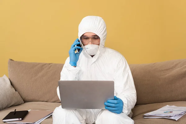 Freelancer en traje de hazmat, guantes de látex y máscara médica hablando en smartphone y usando laptop en sofá - foto de stock