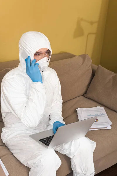 Фрилансер в защитном костюме и медицинской маске разговаривает на смартфоне, используя ноутбук рядом с документами на диване — стоковое фото
