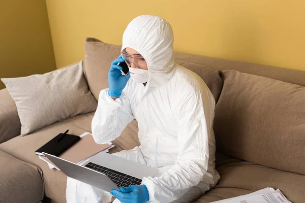 Freelancer em terno hazmat e luvas de látex falando no smartphone e usando laptop no sofá na sala de estar — Fotografia de Stock