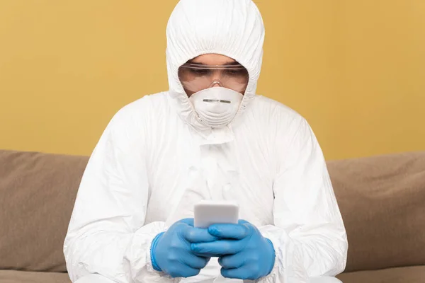 Enfoque selectivo del hombre en traje de materiales peligrosos, guantes de látex y máscara médica utilizando el teléfono inteligente en el sofá en casa - foto de stock