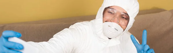 Панорамный снимок человека в защитном костюме и медицинской маске, показывающий знак мира и делающий селфи со смартфоном дома — стоковое фото