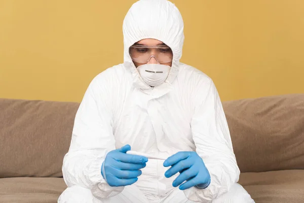 Hombre con traje de materiales peligrosos y máscara médica que sostiene el termómetro en el sofá - foto de stock