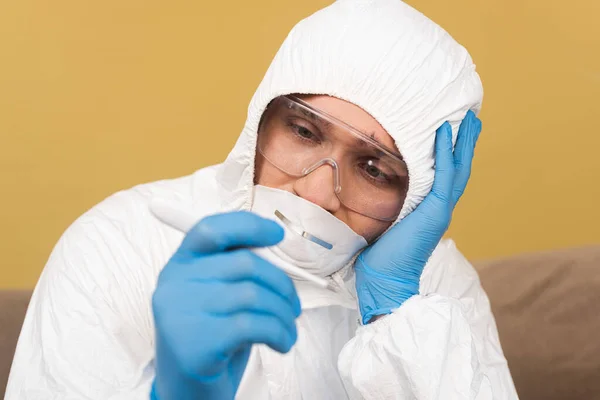 Enfoque selectivo del hombre molesto en traje de materiales peligrosos y guantes de látex que sostienen el termómetro en casa - foto de stock
